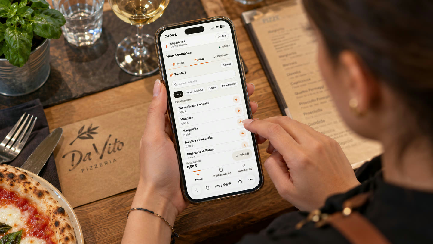 Uno smartphone in mano a chi serve, con l'app del personale di Judgy aperta su «Nuova comanda»: il tavolo 1 è scelto e sotto compaiono le pizze del menù, ognuna con il suo prezzo e il pulsante per aggiungerla. Sul tavolo di legno, una pizza, un calice di vino e il menù cartaceo